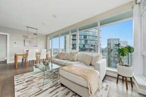 A modern living room with a glass coffee table, a white sofa, and a large window offering a city view. - Sky-High Luxury in Vibrant Yaletown Hub WaterViews (Vancouver)