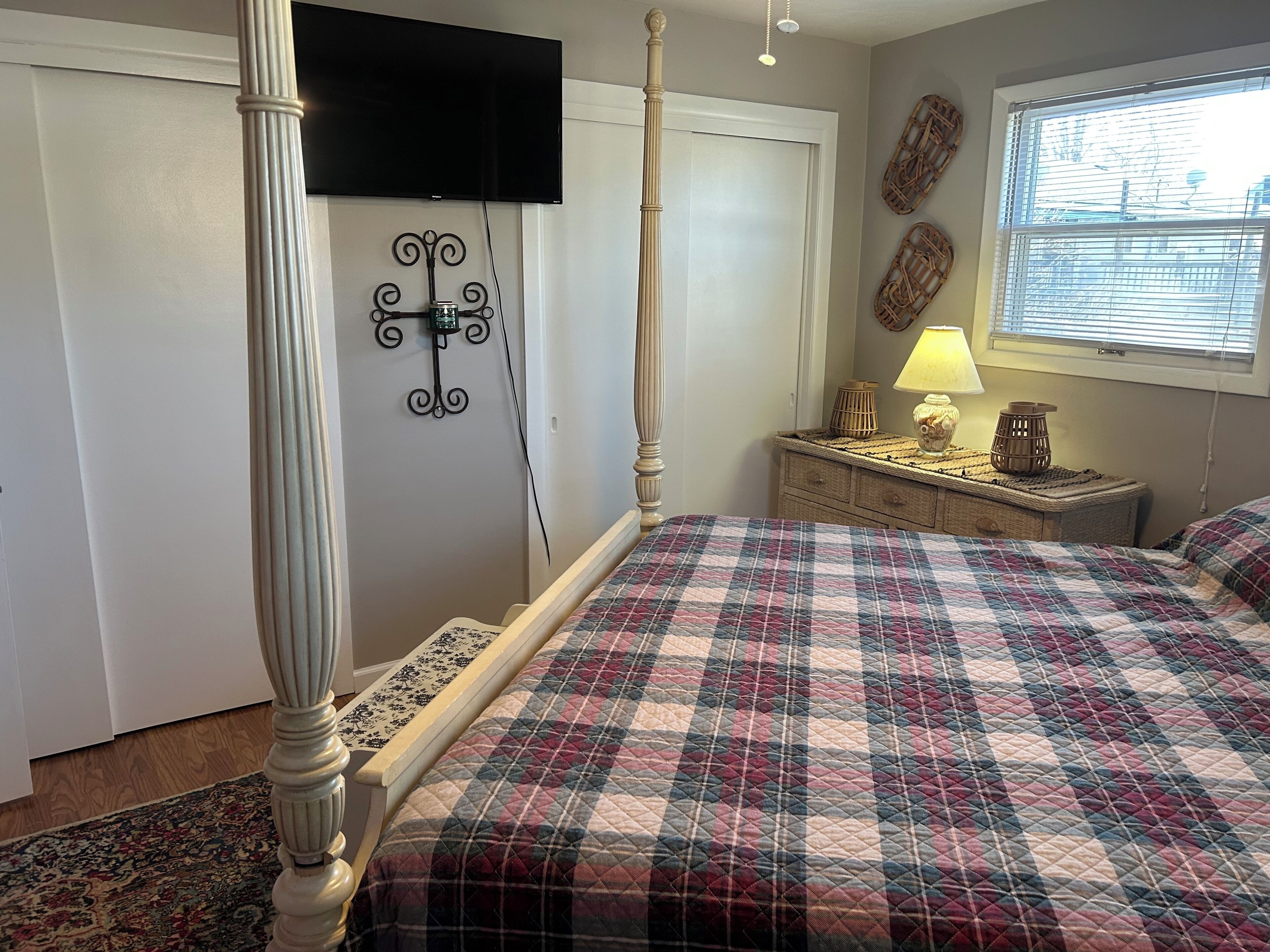 A bedroom with a four-poster bed, a television mounted on the wall, a bedside table with a lamp, and a window with blinds.