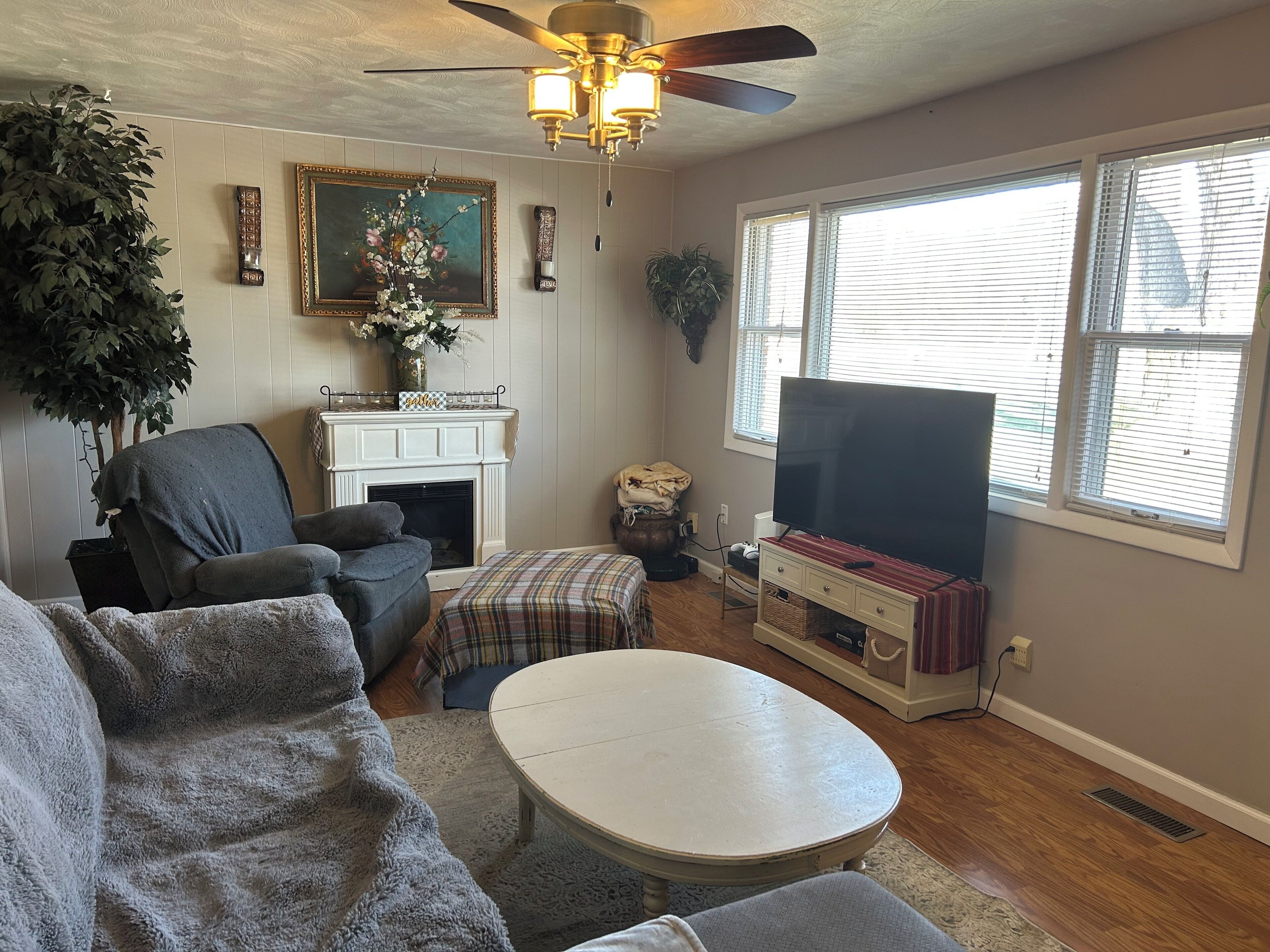 A living room with a fireplace, a television, a ceiling fan, and a large window.