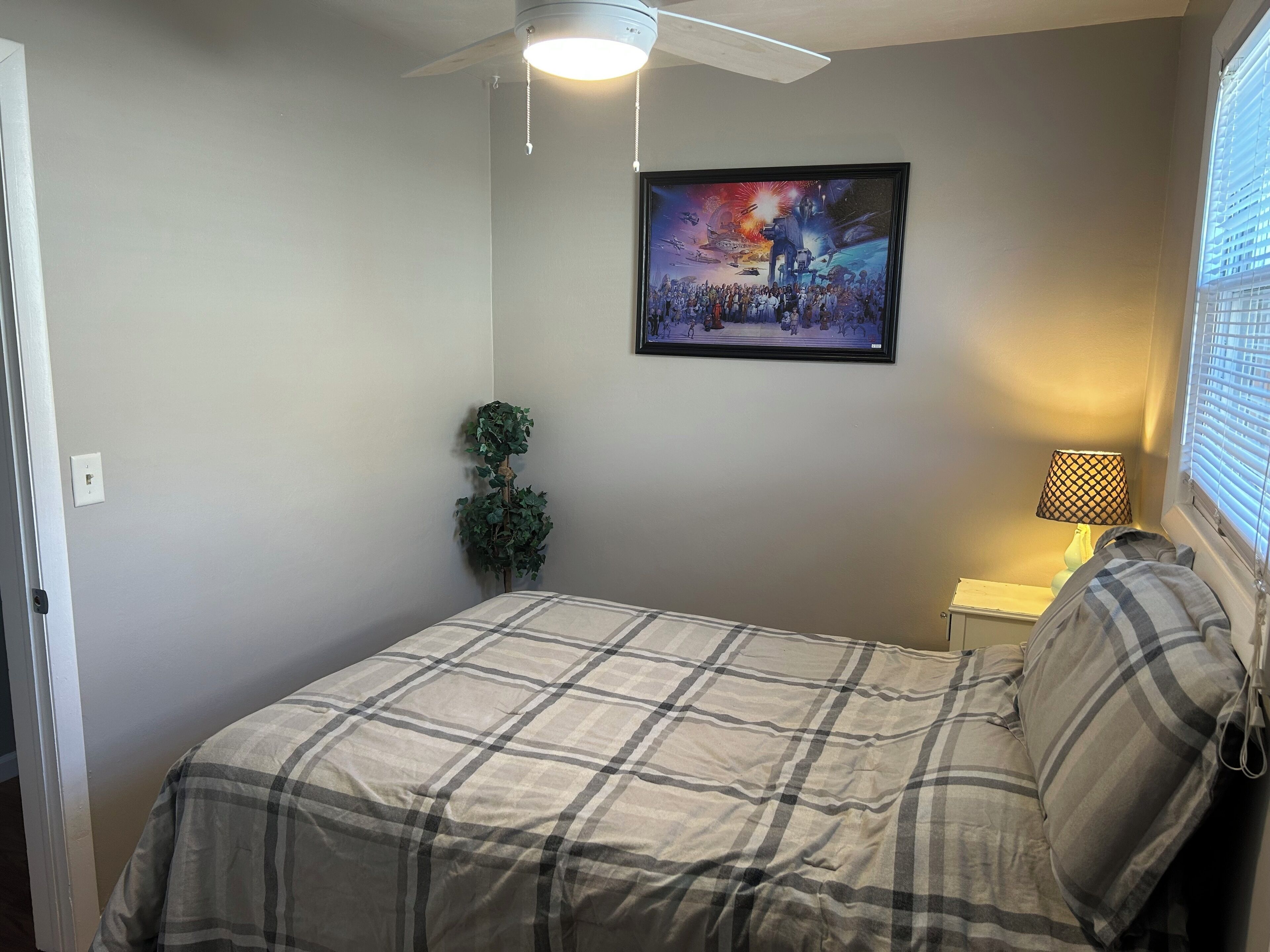 Une chambre à coucher comprenant un lit, un ventilateur de plafond, un tableau accroché au mur, une lampe de chevet et une fenêtre avec des stores.