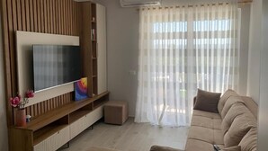 Un salon doté d’un téléviseur à écran plat, d’un mur lambrissé en bois, d’un canapé beige et d’une fenêtre ornée de rideaux légers.