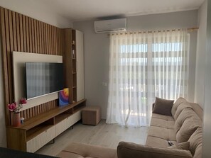 A living room with a flat-screen TV, wooden panel wall, beige sofa, and a window with sheer curtains. - Fully furnished, New stylish One Bedroom apartment - 120 meters from the beach. (Golem)