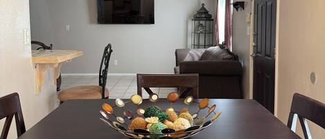 Comedor con mesa de madera oscura, sillas, una lámpara de araña, ventilador de techo y un televisor montado en la pared.