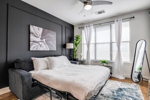 Een moderne slaapkamer met een bed, een grijze bank, een plafondventilator en een groot raam met gordijnen.