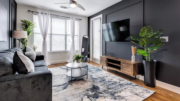 Un salon moderne avec un canapé gris, une table basse ronde, un téléviseur fixé au mur et un ventilateur de plafond.