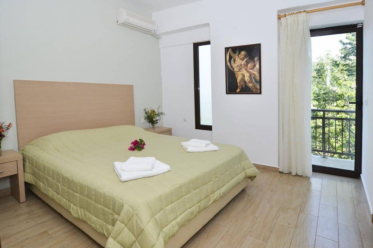 A hotel room with a large bed, a nightstand, a window with a view of greenery, and a painting on the wall.
