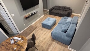 Een moderne woonkamer met een grijze bank, een blauwe poef, een houten salontafel en een flatscreen televisie die aan de muur is bevestigd.