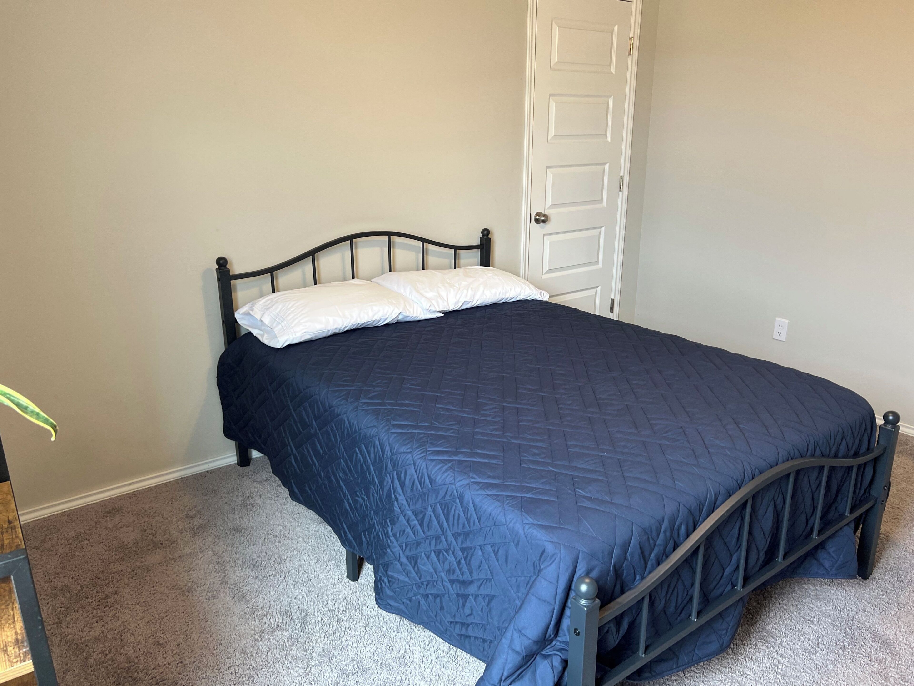 Una cama individual con colcha azul oscuro y almohadas blancas en una habitación con paredes color beige y una puerta blanca.