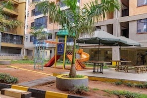 A playground with slides and climbing structures, surrounded by a landscaped area with benches and a palm tree. - Kaisa Gardens (Nairobi)
