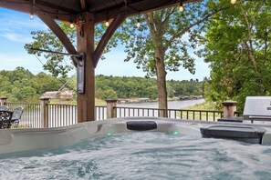 Un jacuzzi con vista a un lago y árboles, terraza de madera y mobiliario exterior.