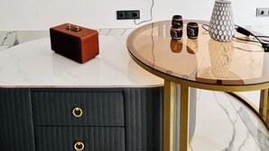 Ein modernes Wohnzimmer mit einem Wand-Fernseher, einem runden Glastisch mit weißer Marmorplatte, einem kleinen roten Lautsprecher, einer weißen Lampe mit geometrischem Fuß und einem dunklen Schrank mit zwei Schubladen.