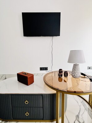 A modern living room with a flat-screen TV mounted on the wall, a round glass coffee table with a white marble top, a small red speaker, a white lamp with a geometric base, and a dark-colored cabinet with two drawers. - Mindelo's Brand New, Modern and Cozy: Family-Friendly Central Apartment for You! (Mindelo)