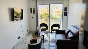 Ein modernes Wohnzimmer mit Marmorboden, einer Ledersofa, einem an der Wand montierten Fernseher und Blick auf eine Hügellandschaft durch große Fenster.