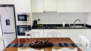 Eine moderne Küche mit weißen Schränken, einem eingebauten Ofen, einer Mikrowelle und einem Gasherd. Es gibt einen Esstisch mit weißen Stühlen und eine Kaffeemaschine.