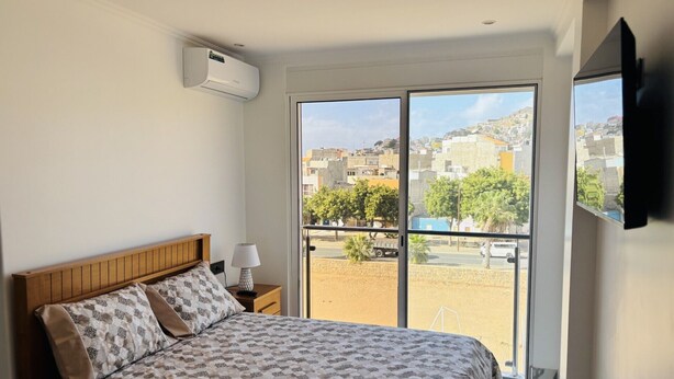 Ein Schlafzimmer mit einem Bett, einem Nachttisch, einer Lampe und einem Fernseher. Es gibt ein Fenster mit Blick auf Gebäude und Bäume.