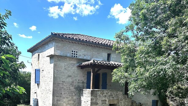 A stone building with a tiled roof, a small porch, and a window with blue shutters.