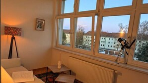 A cozy living room with a white sofa, a small table, a colorful rug, and a telescope on a tripod by a large window. - Children welcome! Spacious family apartment 5 minutes from the center (Lübben (Spreewald))