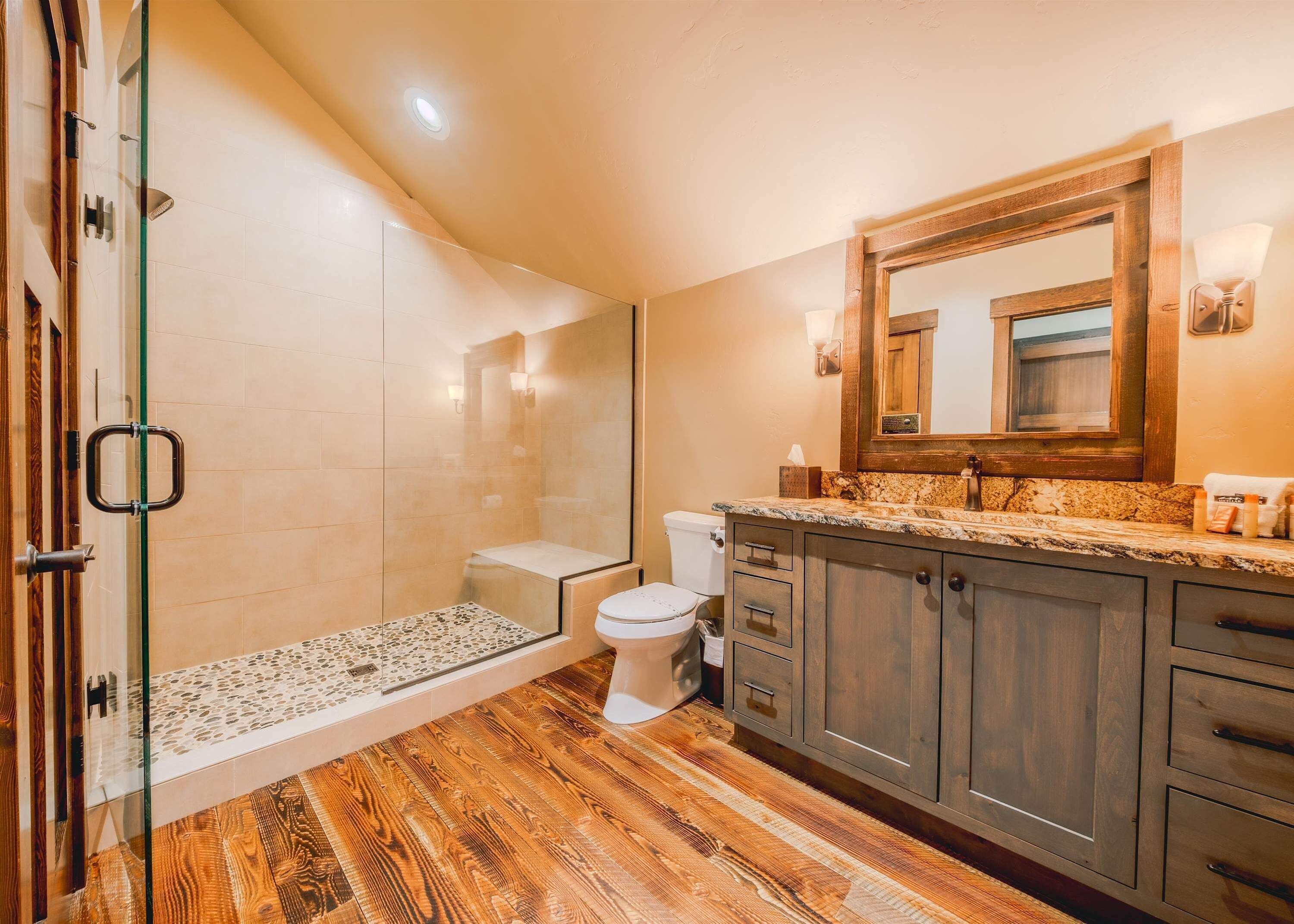 A modern bathroom with a glass-enclosed shower, wooden flooring, a marble countertop, and wooden cabinets.