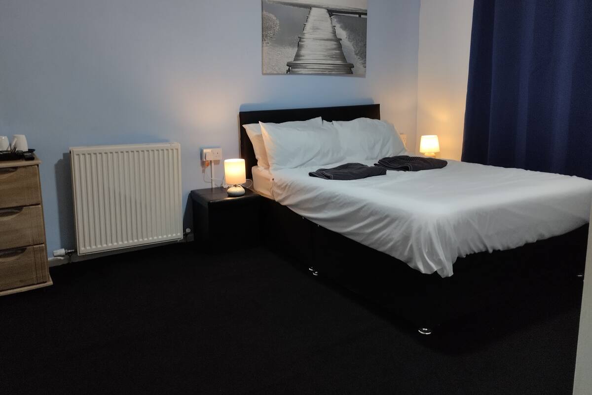 A neatly made bed with white linens, two bedside lamps, a radiator, and a wooden dresser.