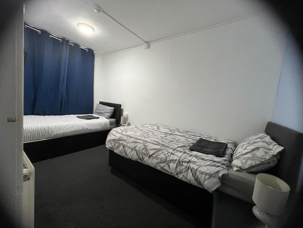 Una habitación pequeña y moderna con dos camas individuales, una mesita de noche con lámpara y una cortina en la ventana.
