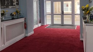 Une pièce spacieuse avec un tapis rouge, une console blanche ornée de fleurs et une grande fenêtre donnant sur la rue.
