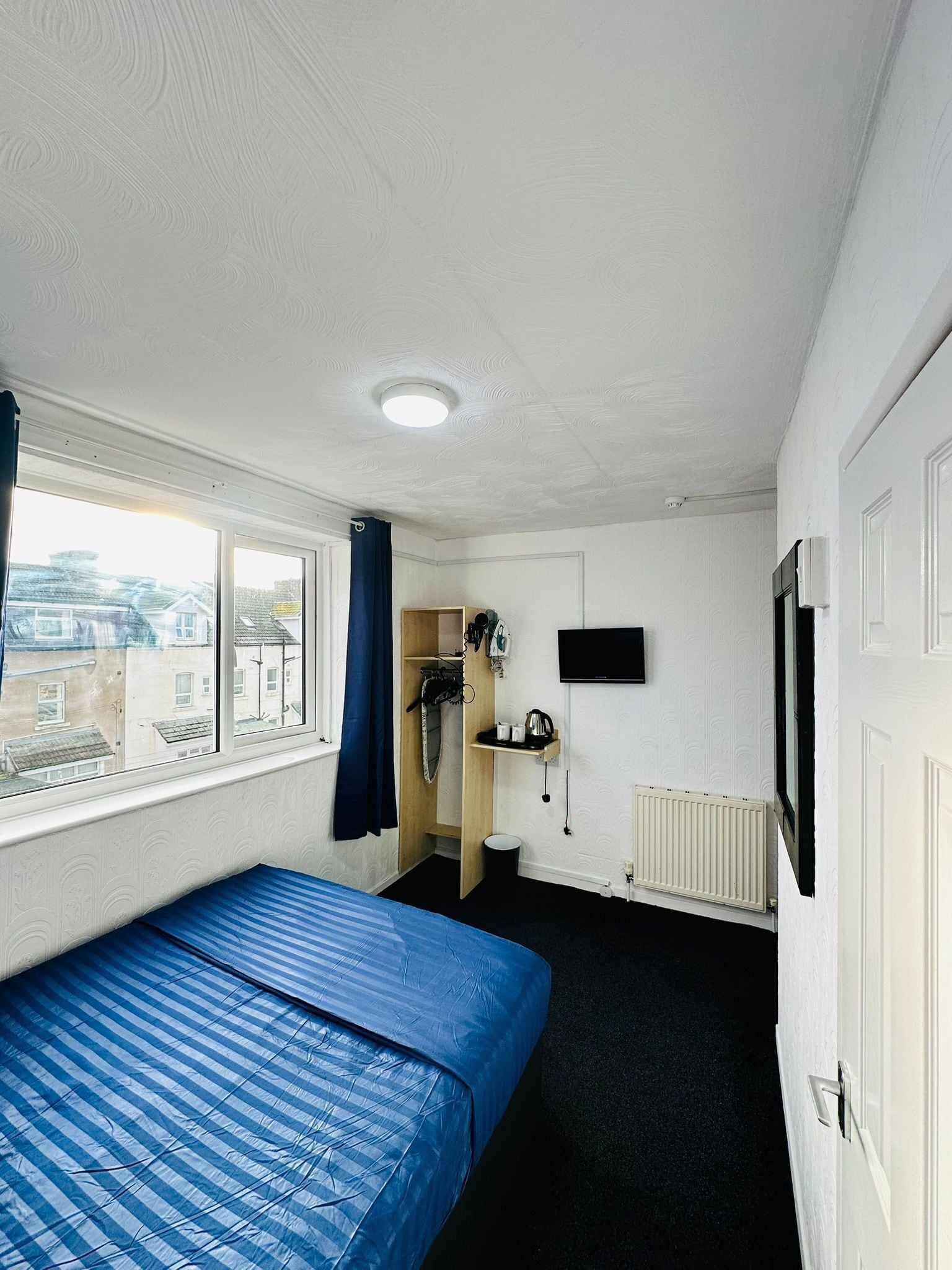Une chambre avec un lit simple, un téléviseur fixé au mur, un radiateur et une fenêtre donnant sur les bâtiments environnants.