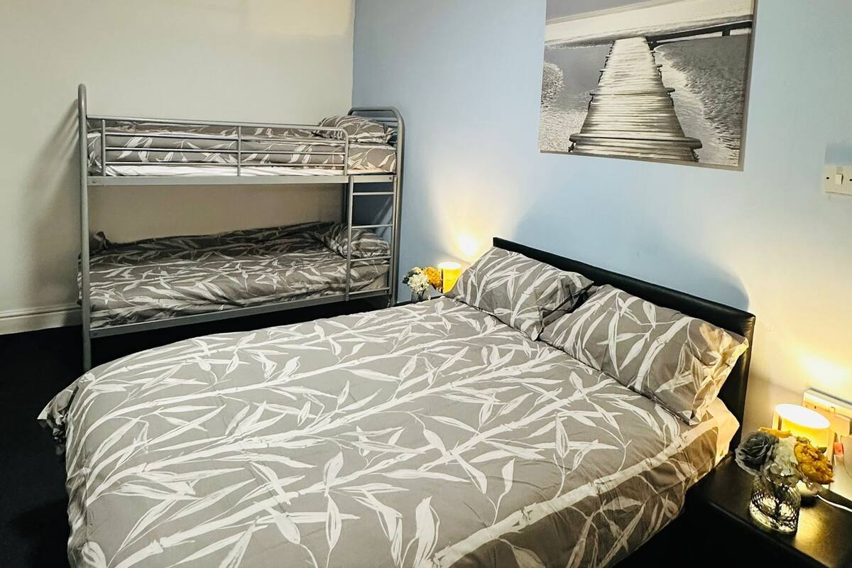A bedroom with bunk beds, a bed with a leaf-patterned bedspread, a wall-mounted picture of a pier, and bedside lamps.