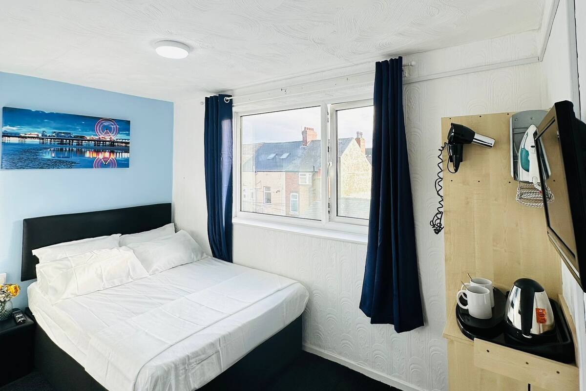A hotel room with a bed, a window with curtains, a wall-mounted phone, a coffee maker, and a painting of a cityscape.