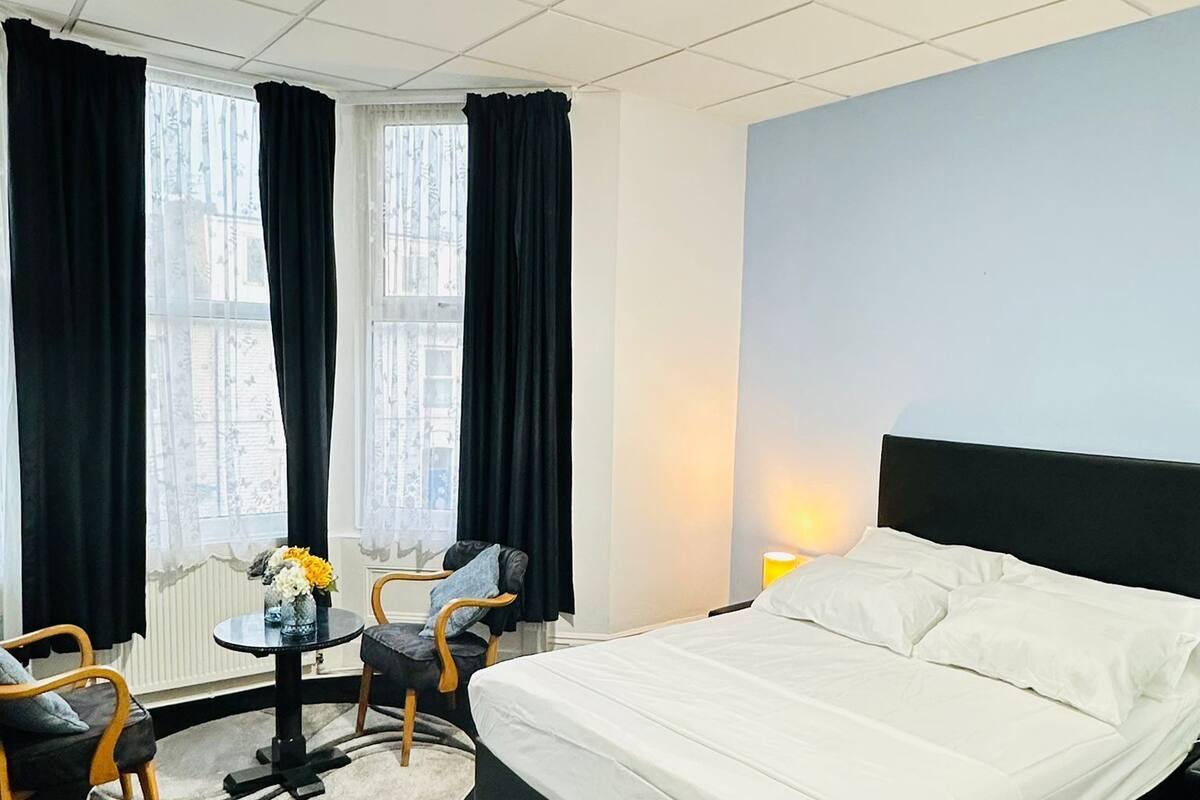 A hotel room with a bed, a round table with chairs, a window with curtains, and a nightstand with a lamp.