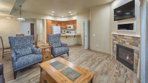 A spacious living room with a fireplace, two patterned armchairs, a wooden coffee table, and a kitchen in the background. - 1 Bedroom Villa, Heated Outdoor Pool and Hotsprings - Gorgeous mountain views (Genoa)