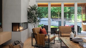 Ein modernes Wohnzimmer mit Kamin, bequemen Sitzgelegenheiten, einem großen Fenster und Blick ins Freie.