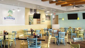 L’intérieur d’un restaurant, avec de nombreuses tables et chaises, un ventilateur de plafond et des décorations murales, dont une enseigne indiquant « Restaurant et bar Breezes ».