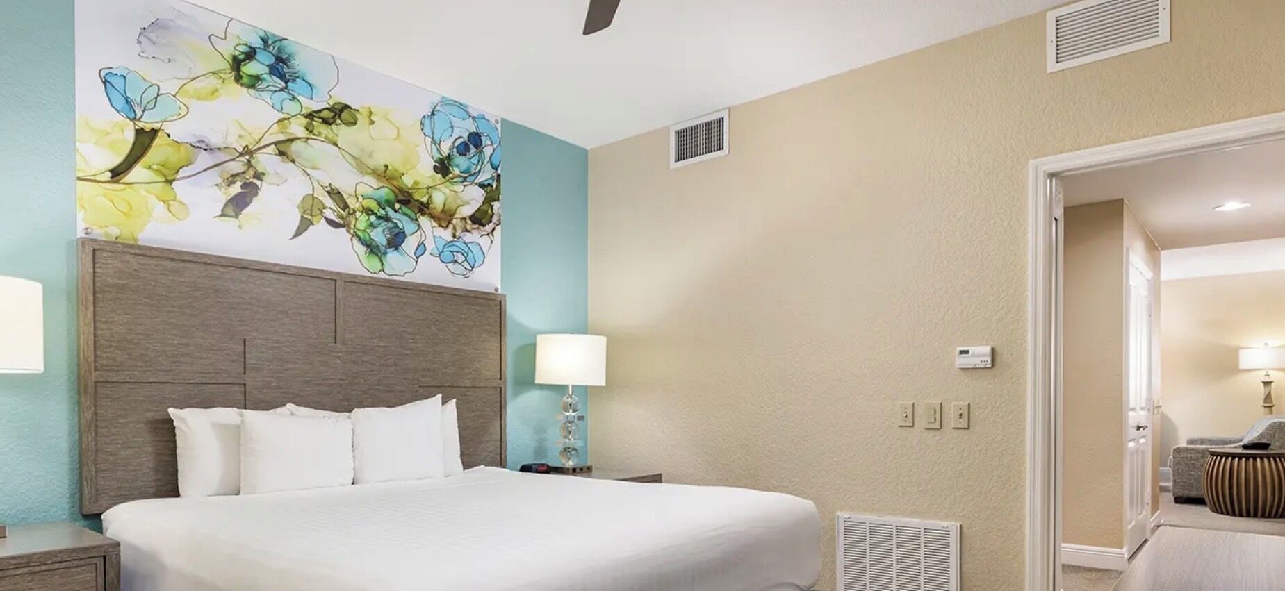 A hotel room with a large bed, a headboard, a wall-mounted lamp, and a floral artwork.