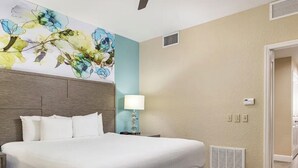 Une chambre d’hôtel avec un grand lit, une tête de lit, une lampe fixée au mur et une œuvre d’art à motifs floraux.