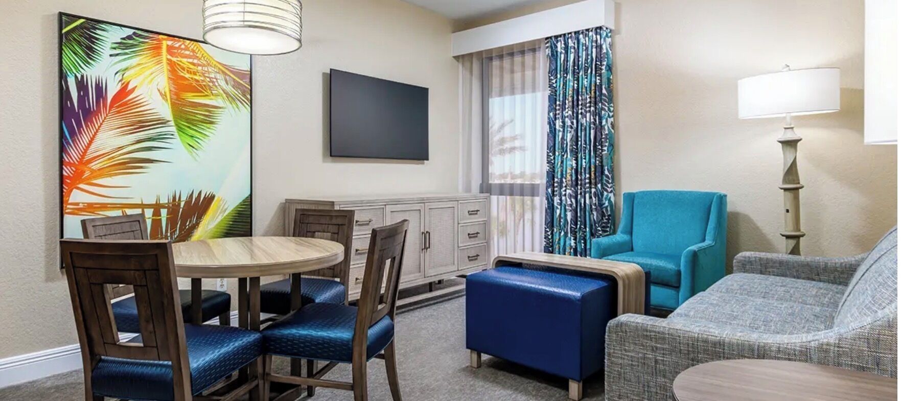 A hotel room with a dining table, chairs, a sofa, a blue ottoman, a TV, a lamp, and a window with curtains.