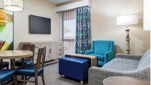Une chambre d’hôtel comprenant une table à manger, des chaises, un canapé, un pouf bleu, une télévision, une lampe et une fenêtre avec des rideaux.