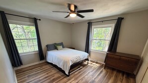 Un dormitorio con cama, ventilador de techo, cómoda y dos ventanas con cortinas.