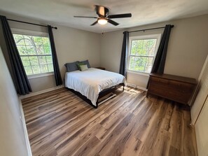 A bedroom with a bed, a ceiling fan, a dresser, and two windows with curtains.