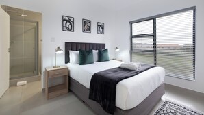 Une chambre moderne comprenant un lit, des tables de chevet, une commode, une douche et une fenêtre avec des stores.