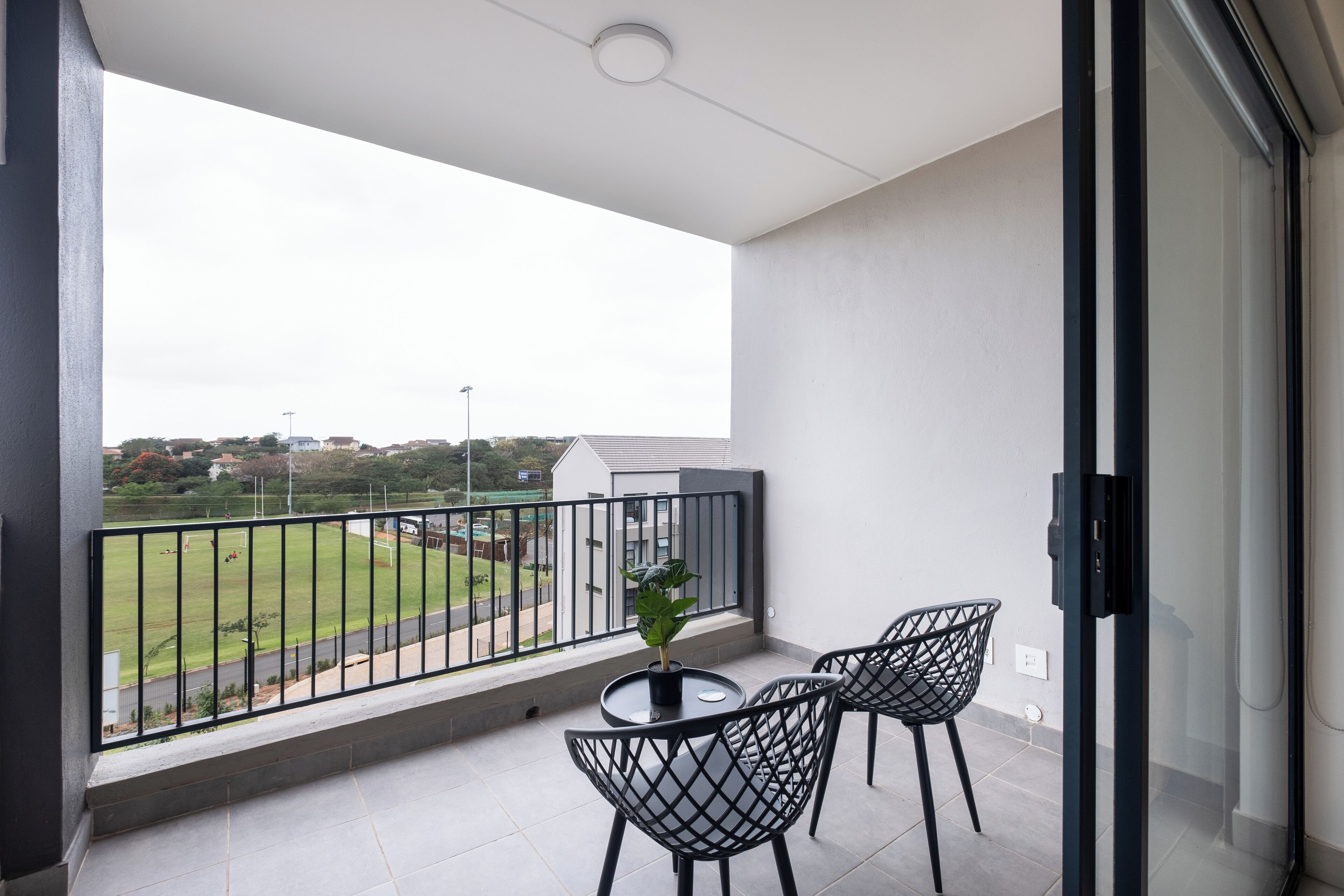 A balcony with a view of a sports field, a potted plant, and two chairs.