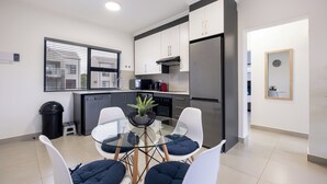 Une cuisine moderne avec des meubles blancs, un réfrigérateur en acier inoxydable et une table à manger en verre agrémentée de coussins bleus.