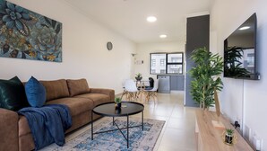 Un salon moderne avec un canapé marron, une table basse et un tapis bleu. On aperçoit une cuisine à l’arrière-plan, avec des meubles blancs et une fenêtre.