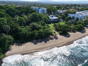 An aerial view of a coastal area with a sandy beach, ocean waves, and residential buildings surrounded by lush greenery.