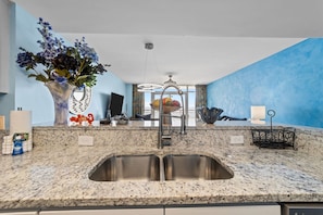 A kitchen with granite countertops, stainless steel sinks, a vase with flowers, and a view of the sea through large windows. - COSTAL VIBES/GREAT LOCATION (Myrtle Beach)