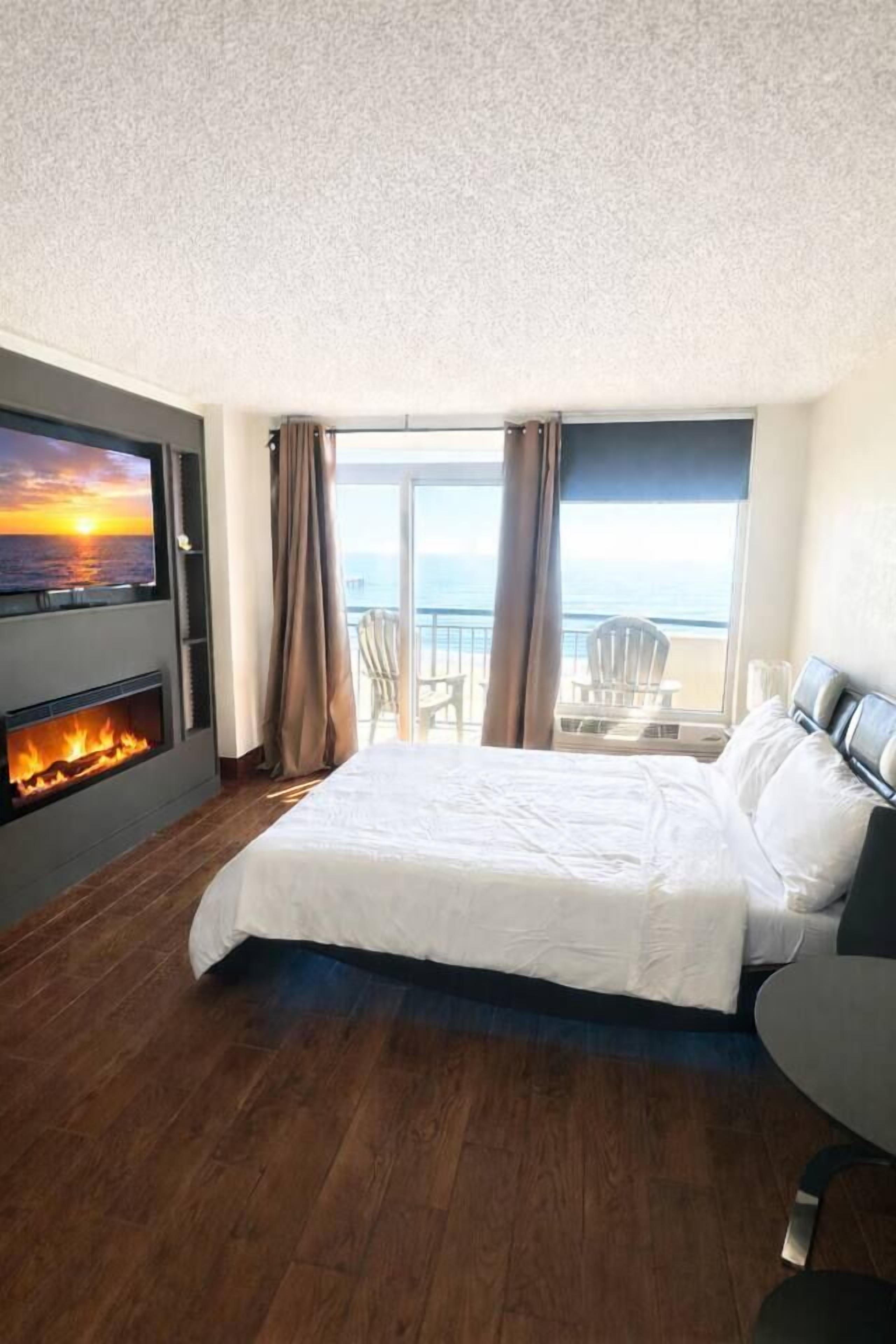 A hotel room with a large bed, a fireplace, a TV mounted on the wall, and a balcony with a view of the sea.