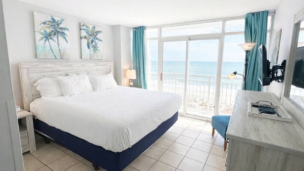 Habitación de hotel con una cama grande, un escritorio con lámpara y vista a la playa.
