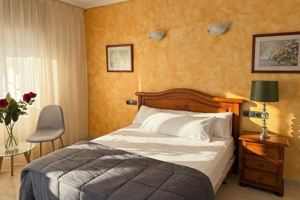 Habitación de hotel con una cama, una silla, una mesita de noche con lámpara y un florero con flores.