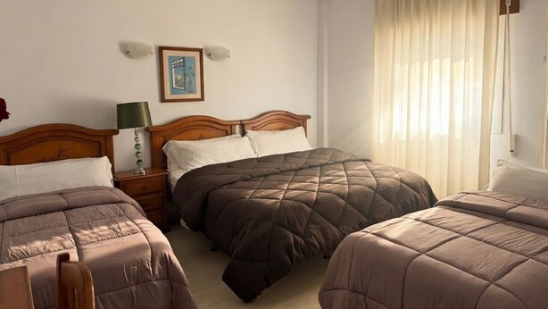 Een hotelkamer met twee bedden, een nachtkastje met lamp, een schilderij aan de muur en een raam met gordijnen.