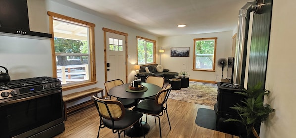 A compact kitchen with a stove, dining table, chairs, and a seating area with a couch and lamp. - American Alps Cabin-Beach, Riverfront, EV charger, 2B/1Ba, North Cascade, Diablo (Marblemount)
