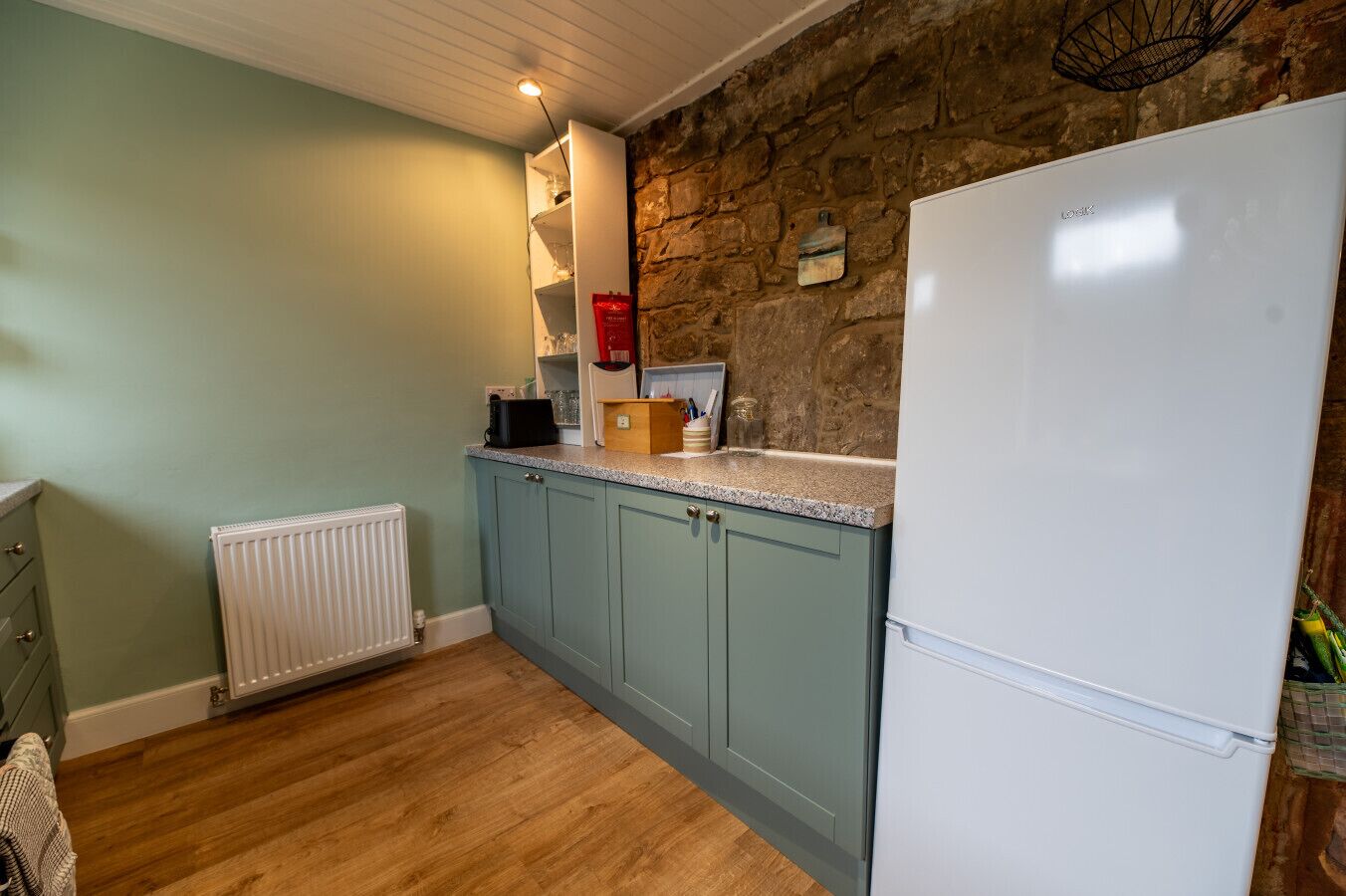 A kitchen with light blue cabinets, a white refrigerator, a stone wall, and a wooden floor.
